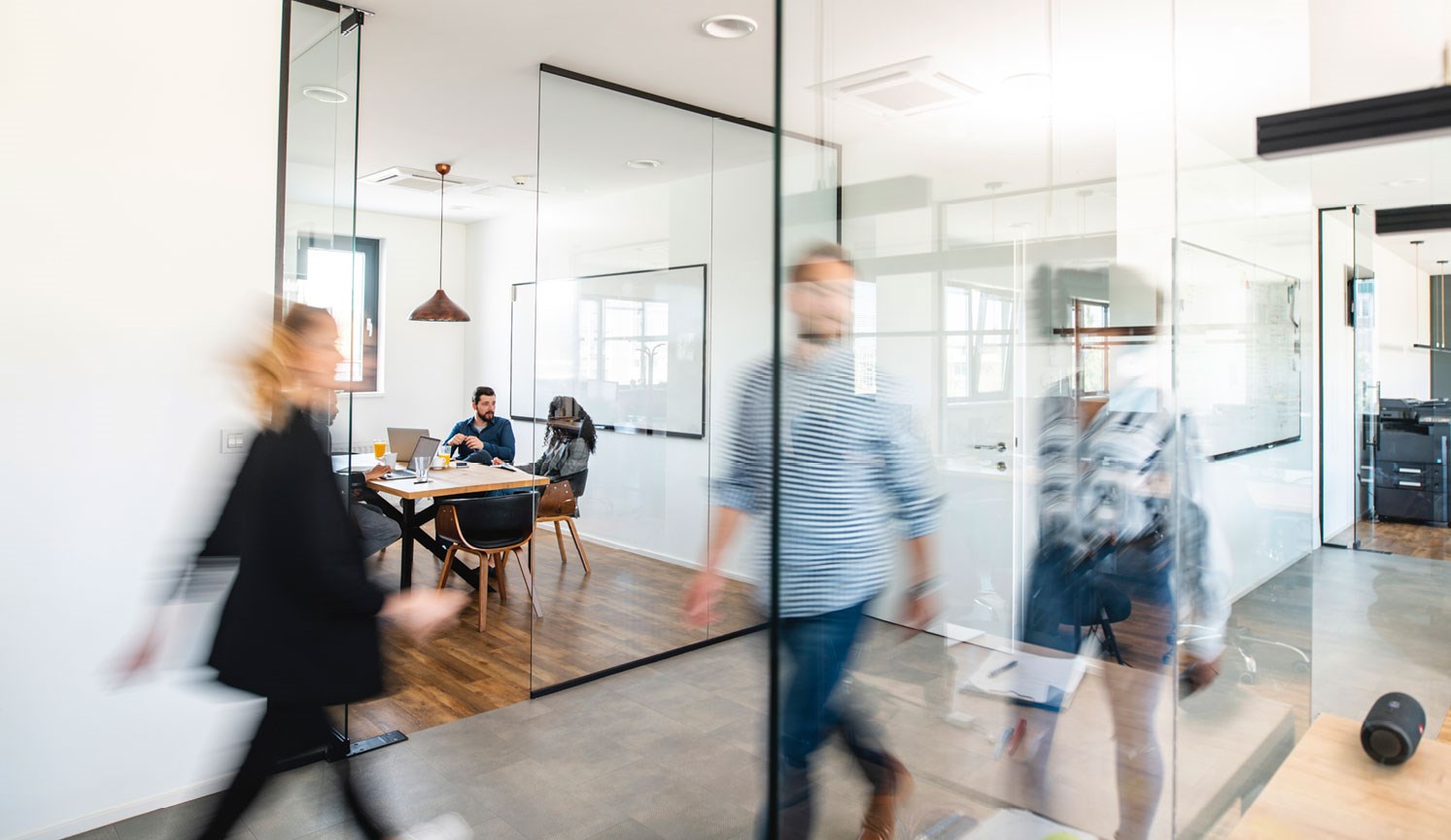 busy office hallway in foreground, people in meeting room in background